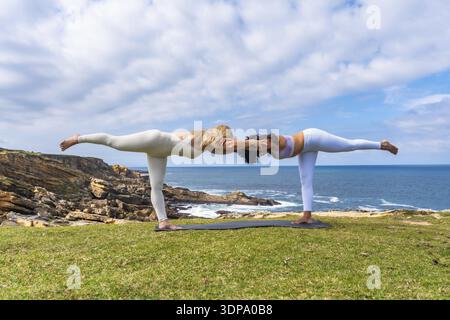 Two women practicing acro yoga outdoors on a grassy cliff overlooking the ocean, holding a deep warrior pose together, symbolizing balance, teamwork, Stock Photo