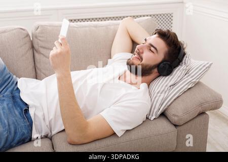 Young Man Relaxes on Sofa While Enjoying Music on His Smartphone in Living Room Stock Photo