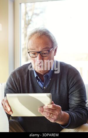 Glasses, surprise and tablet with senior man in living room of home for notification or reaction. App, reading and wow with old person in apartment Stock Photo