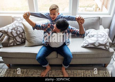 Happy, dad and kid with piggyback on sofa for bonding together, fantasy adventure and weekend fun. Above, father and carry child in home for flying Stock Photo
