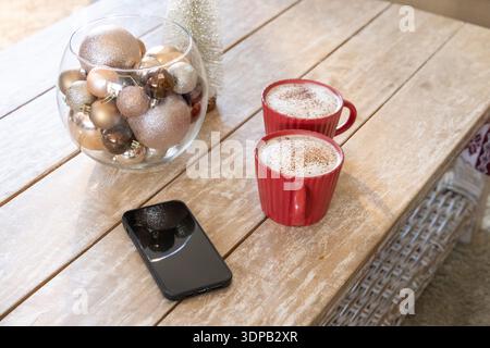 Living room coffee table showing two red mugs frothing with cocoa, metallic baubles, smartphone Stock Photo