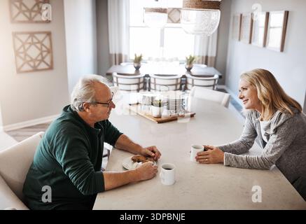 Elderly couple, morning and breakfast in kitchen with coffee for conversation or discussion in home. Senior, man and woman eating with smile, caffeine Stock Photo