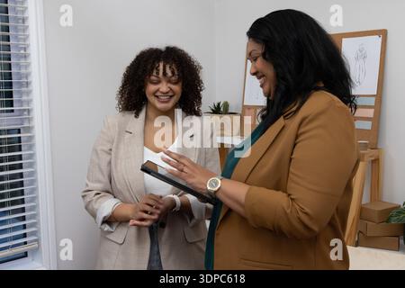 Two female coworkers wearing blazers collaborating in design studio with tablet and sketches Stock Photo