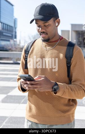 Adult African American man standing in sunlit plaza holding phone, wearing cap, backpack, earbuds Stock Photo