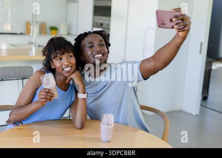 African American couple taking selfie at round table in kitchen, holding pink smoothies, smartphone Stock Photo