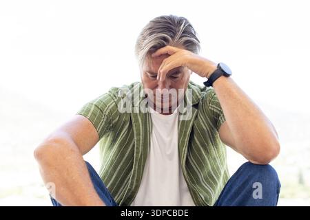 Mature man sitting, bowing head, touching forehead on balcony in green shirt and left-wrist watch Stock Photo