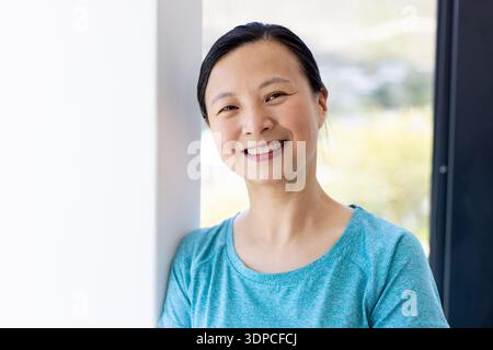 Asian woman leaning against white wall by large window, smiling and facing camera in teal top Stock Photo