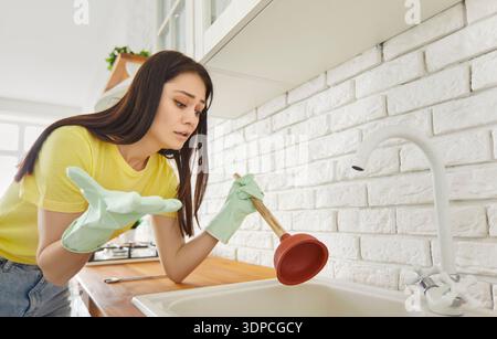 Woman wearing rubber gloves holding plunger stands in cozy kitchen beside clogged sink Stock Photo