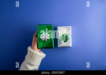 First person top view photo of female hands holding two shiny green and silver gift boxes with bow on isolated blue background Stock Photo