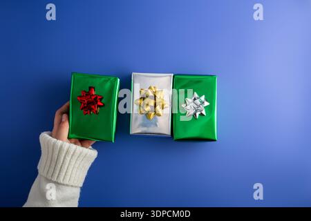 First person top view photo of female hands holding three shiny green and silver gift boxes with bow on isolated blue background Stock Photo