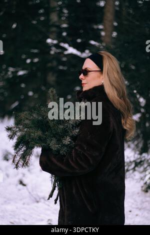 Woman with long blonde hair wearing sunglasses and a dark fur coat holds a bundle of evergreen branches  Stock Photo