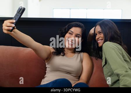 Two female friends sitting close on rust-colored couch at home, holding smartphone and smiling Stock Photo