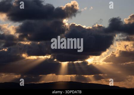 Crepuscular rays of sun breaking through dark clouds over a dark hill silhouette Stock Photo