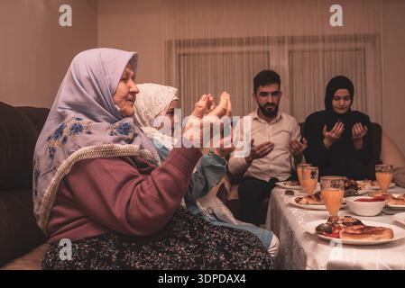 Muslim Family Praying Together Making Dua To Break Their Fast During Ramadan Meal At Home Celebrating Iftar Dinner With Traditional Food And Drinks. Stock Photo