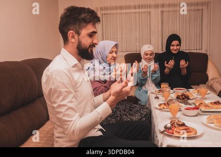 Muslim Family Praying Together Making Dua To Break Their Fast During Ramadan Meal At Home Celebrating Iftar Dinner With Traditional Food And Drinks. Stock Photo