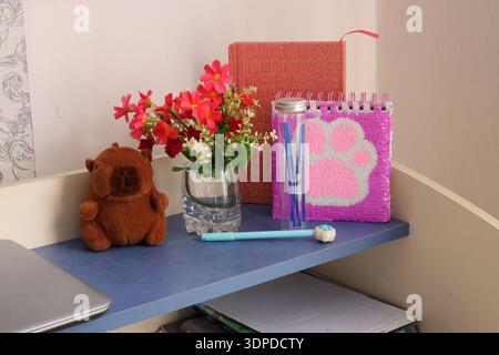 Cozy student or teenager workspace with a cute capybara plush toy, bright pink sequin notebook, a vase of red flowers, and stationery on a blue shelf. Stock Photo