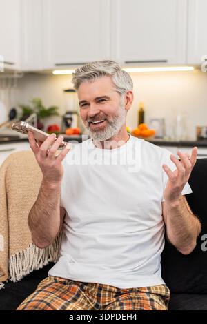 Middle-aged man at home records voice note on smartphone waves hello on speaker and shares news. Mature guy on sofa smiles talks sends chat reply then listens to playback nods and adds a short update. Stock Photo