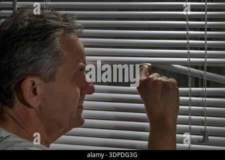 A man monitors what is happening by looking through the slats of a Venetian blind Stock Photo