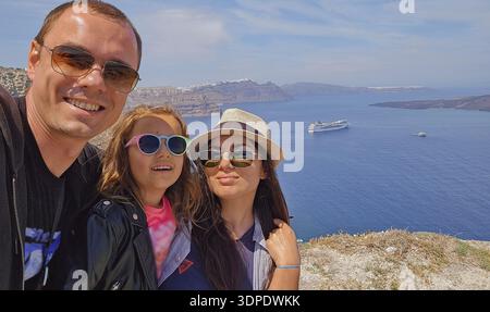 Sunny family selfie with panoramic view of Santorini caldera and Aegean Sea cliffs in bright Mediterranean daylight Stock Photo