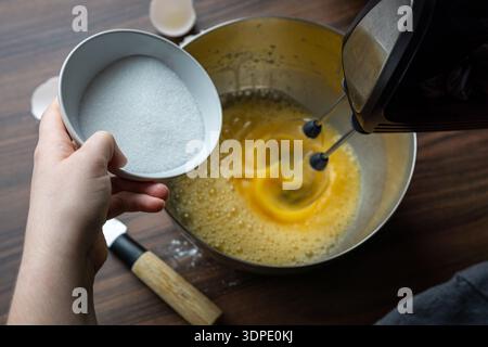 Top view of sugar being added to beaten eggs in a metal bowl while mixing with an electric mixer. Stock Photo