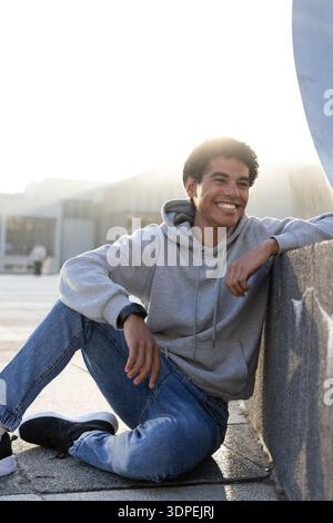 Teenage male sitting on concrete plaza leaning against low wall, smiling, wearing gray hoodie Stock Photo