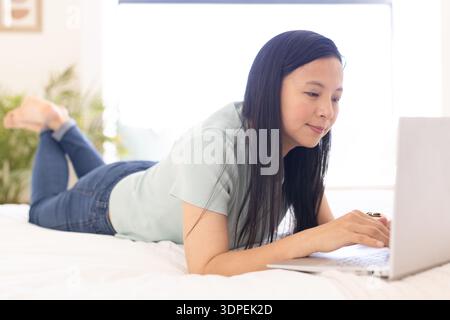 Asian woman lying on stomach on bed, propping on elbows and typing on laptop near window Stock Photo