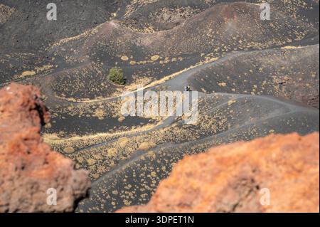 Three hikers walk along a winding trail on the volcanic slopes of Mount Etna, surrounded by dark ash-covered terrain and scattered patches of vegetati Stock Photo
