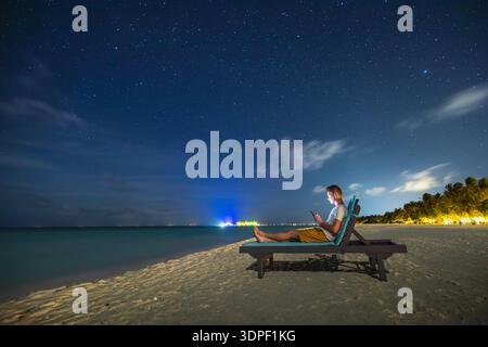 Lonely man lying on sunbed on deserted sandy beach looking at phone under starry night sky. Solitude, digital connection, relaxation, and modern lifes Stock Photo