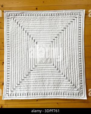 Minimalist handmade quilt wall hanging with concentric square frames in neutral linen tones, featuring a stylized line-drawn female face in the center. Stock Photo