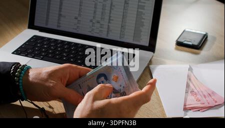 Person counts cash next to a laptop and smartphone on a wooden table during the day in a home office setting Stock Photo