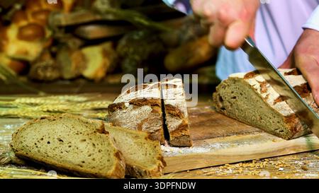 Baker cuts fresh bread on a wooden board in a bakery with various types of bread in the background during daytime Stock Photo
