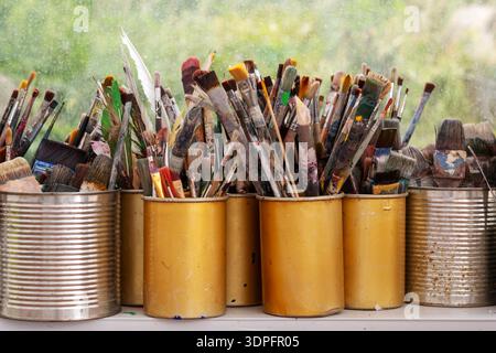Rows of well used paintbrushes stand in yellow and silver cans on a studio windowsill layered with dried color against a green garden view creating an Stock Photo