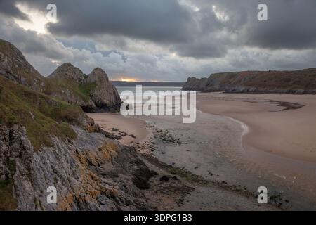 Sunlight glows on distant water beneath dark storm clouds, as tidal channels curve across a broad sandy beach on the Gower Peninsula, Wales. Stock Photo