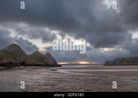 Storm clouds over Three Cliffs Bay with sunset light breaking on the horizon and a tidal stream crossing the sandy beach, Gower Peninsula, Swansea, Wa Stock Photo
