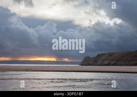 Dramatic storm clouds over Three Cliffs Bay with sunset light on the horizon and a tidal stream crossing the sandy beach, Gower Peninsula, wales Stock Photo