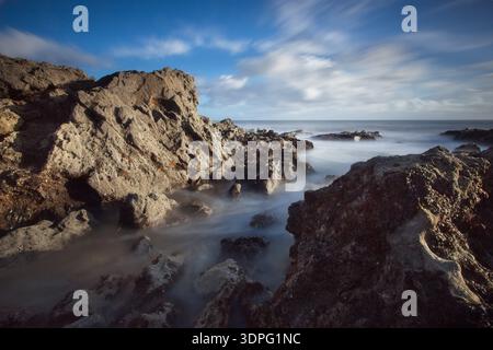 Rock formations and surf nr. Langland, Gower coast in Wales, captured with long exposure for soft water and moving clouds under blue sky. Stock Photo