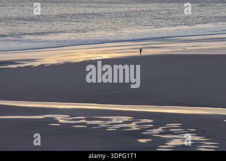 Distant photographer walking along the shoreline as golden light reflects across wet sand and shallow pools. Stock Photo