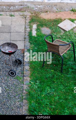 A cozy backyard patio scene with three wicker chairs around a red wooden table, scattered pinecones, a small charcoal grill on gravel and a blue tarp Stock Photo