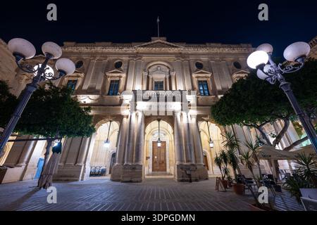Illuminated Neoclassical facade of the National Library of Malta in Valletta, with arched entrance, columns, street lamps and courtyard trees at night Stock Photo