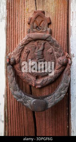 Close-up of a rusty antique cast iron door knocker with ornate relief on a textured red wooden door. Stock Photo