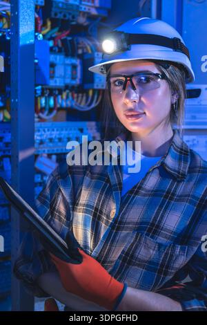 Female electrician with headlamp poses by a switchboard. Tech portrait. Stock Photo