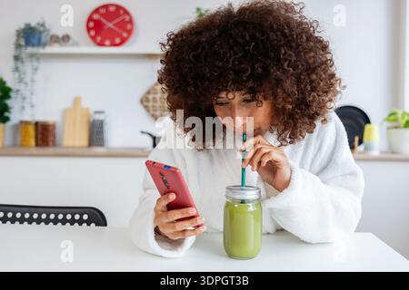 Woman enjoying healthy green smoothie and browsing smartphone in kitchen Stock Photo