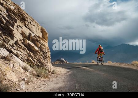 Mountain biker riding on asphalt road near massive jagged rock cliff in Ladakh India featuring white monastery on distant hill under dramatic dark sto Stock Photo