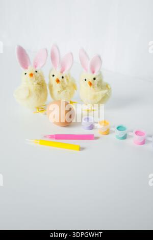 One Easter egg with bright colorful paints, markers and funny small toy chickens with Easter bunny ears on white background Stock Photo