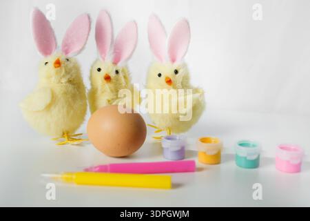 One Easter egg with bright colorful paints, markers and funny small toy chicks with Easter bunny ears on white background Stock Photo