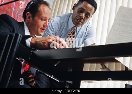Two men are seated at a table in an office. They are focused on documents and engaged in conversation. They appear to be working on a project together Stock Photo