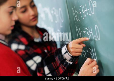 Two students are working together on chemistry problems at the blackboard. They focus on writing and solving equations on a chalkboard. Their expressi Stock Photo
