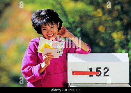 And attractive Asian woman smiles as she stands next to a mailbox reading her mail. She is holding several postcards and letters in her hands. The sur Stock Photo