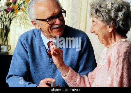 A senior couple smile and interact in a warm indoor space. The man  is handling her engagement ring and proposing. Flowers add color to the scene. Stock Photo