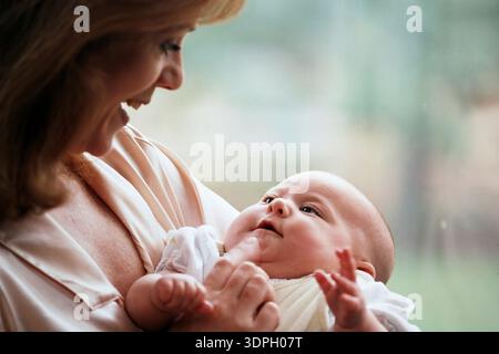 A mother smiles as she holds a baby close. The baby looks back at her with curiosity. They are indoors with natural light shining through a window. Stock Photo